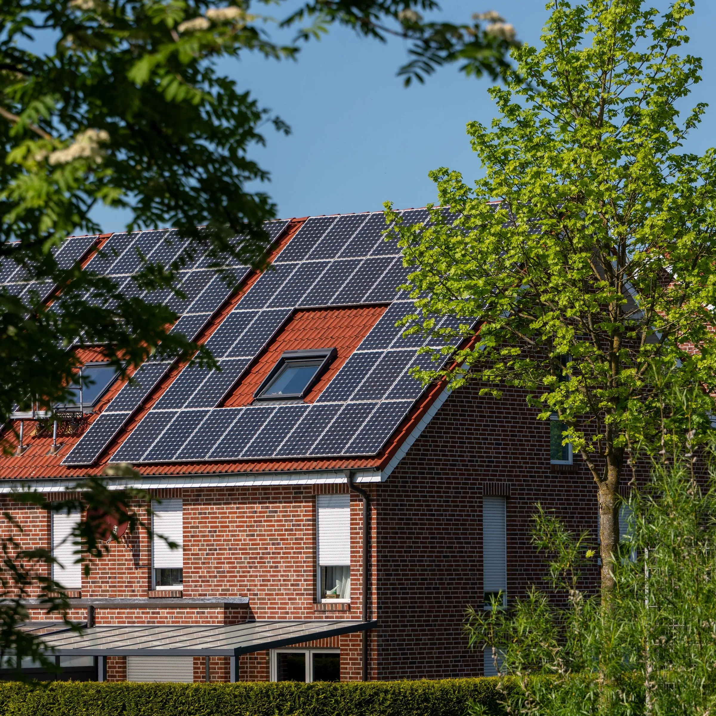 Backsteinhaus mit rotem Satteldach, dessen Dachfläche mit Solarmodulen belegt ist, davor Laubbäume